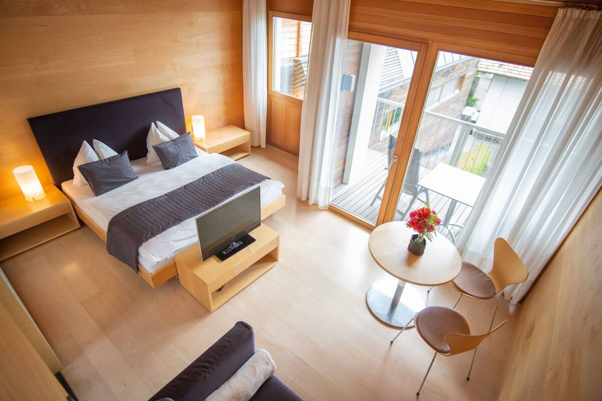 A modern bedroom with a bed, bedside tables, a TV, a small table with chairs, and a balcony with a view.