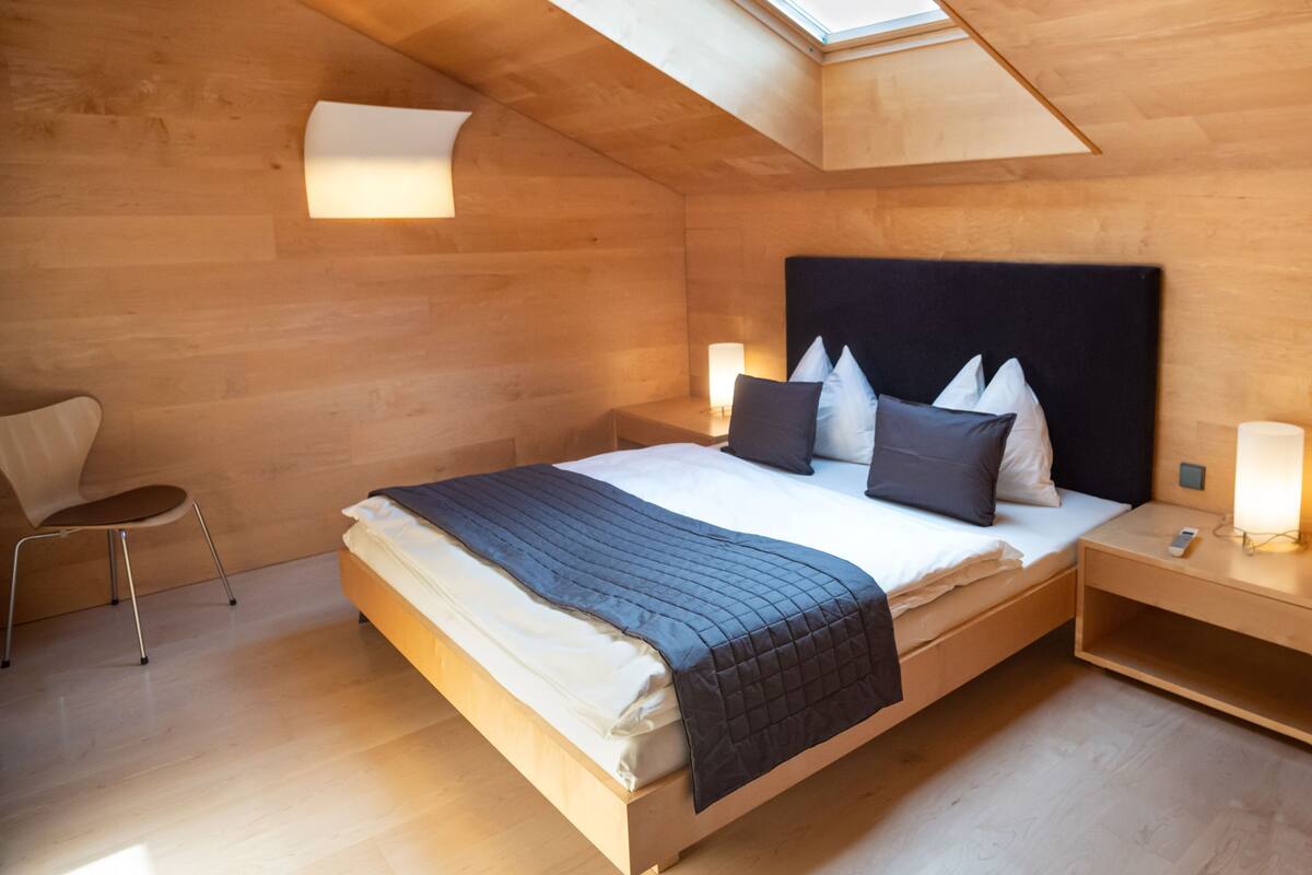 A bedroom with a skylight, a bed with pillows, a bedside table with a lamp, and a chair.