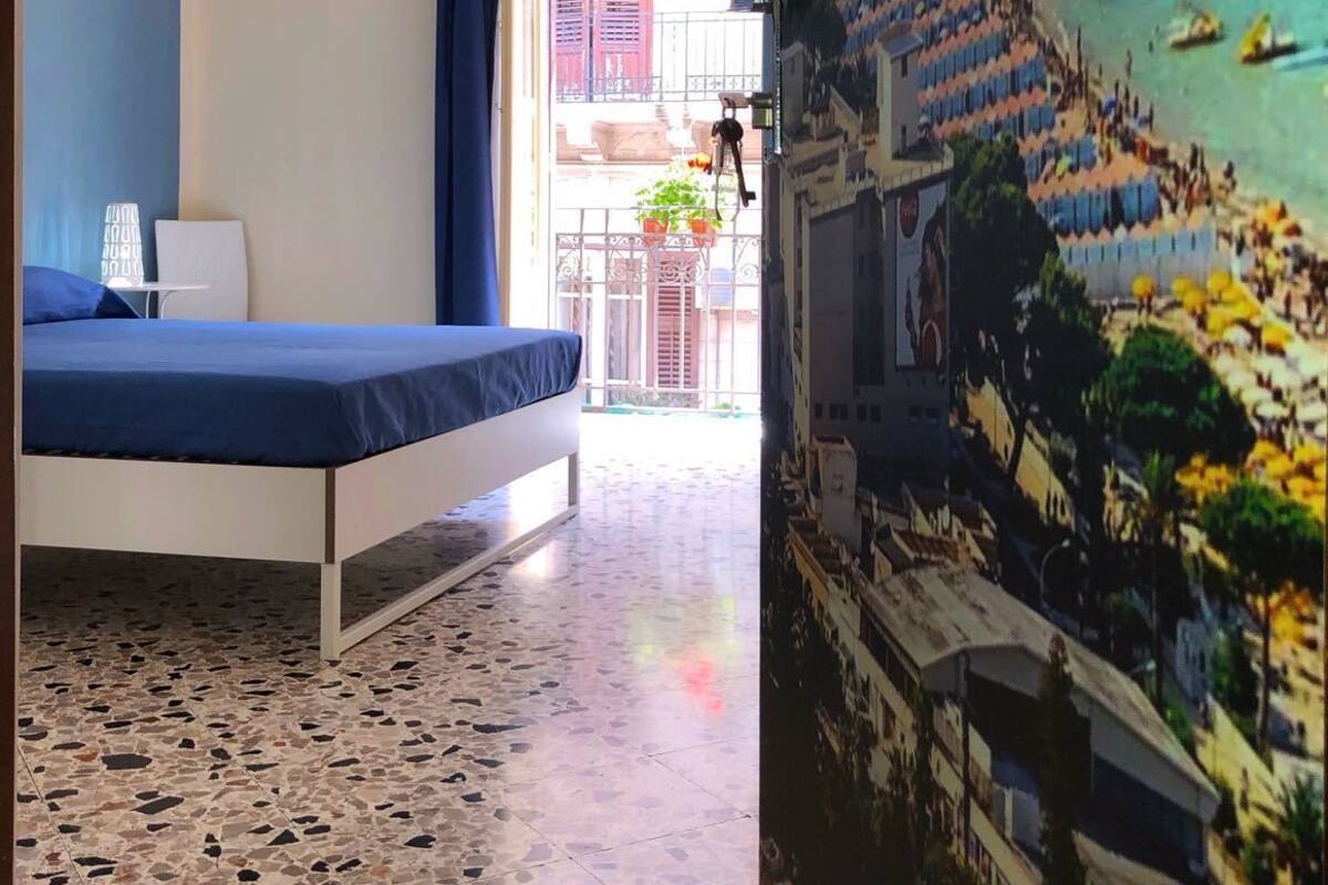 A room with a bed, a window with curtains, a tiled floor, and a view of a beach with people and boats.