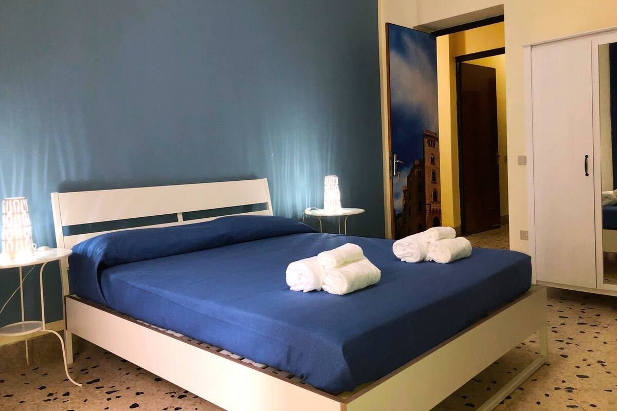 A hotel room with a bed, a small bedside table with a lamp, and a door leading to another room.