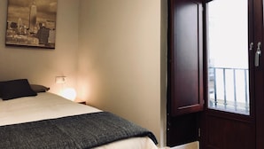A hotel room with a bed, a nightstand with a lamp, a cityscape painting on the wall, and a balcony door.