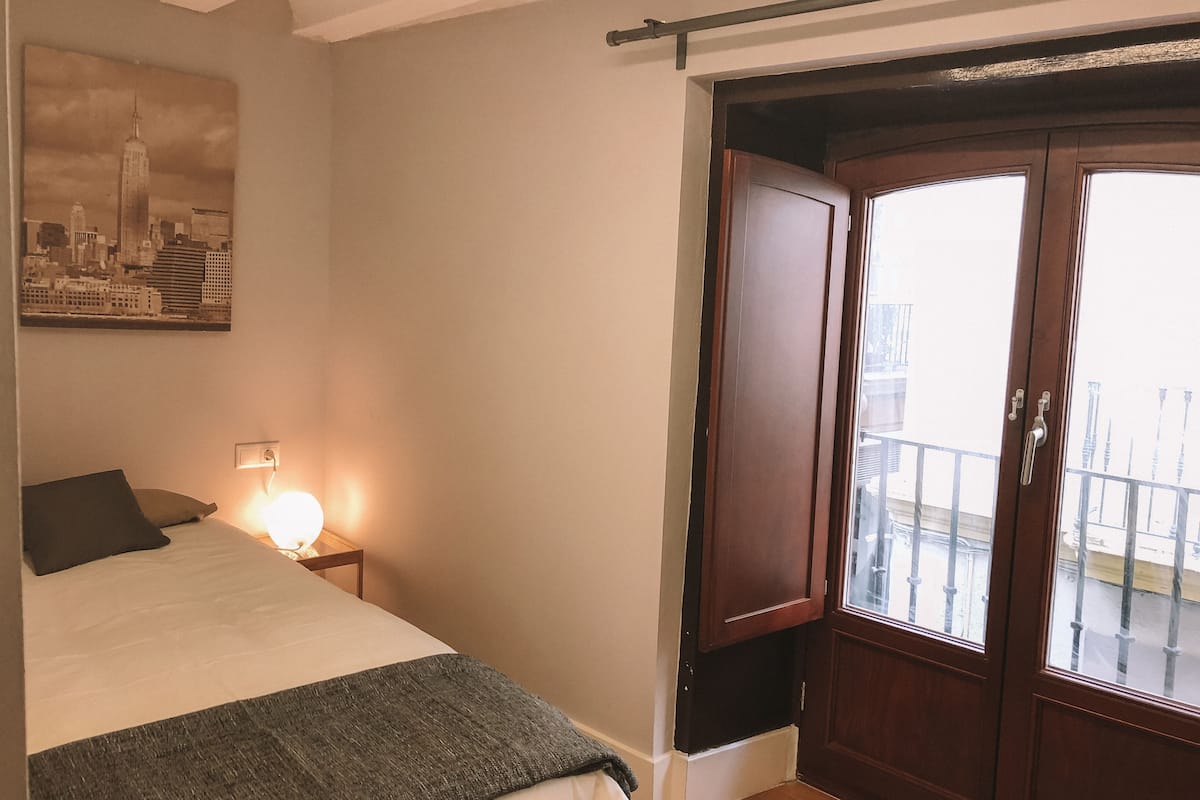 Une chambre à coucher avec un lit, une table de chevet avec une lampe, une porte donnant sur un balcon et un tableau représentant un paysage urbain accroché au mur.