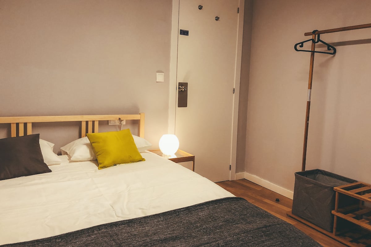 Un lit bien fait, avec une tête de lit en bois, une lampe de chevet et une boîte de rangement grise.