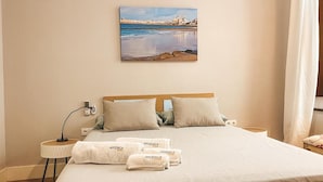 A neatly made bed with a wooden headboard, two pillows, and a blanket. There are towels with the brand 'APODACA' on the bed. A small bedside table with a lamp and a phone charger is visible. A painting of a coastal scene hangs on the wall above the bed.