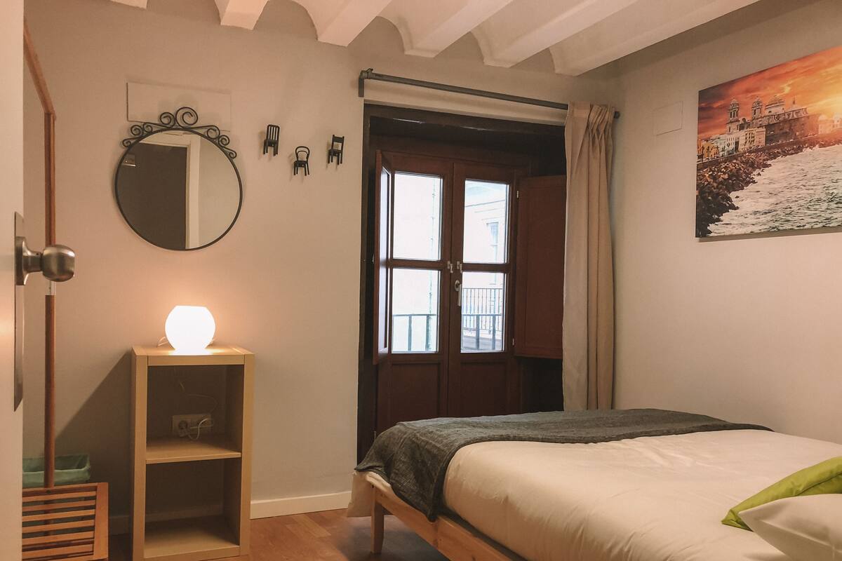 Une chambre à coucher avec un lit, une table de chevet avec une lampe, un miroir et une fenêtre avec des rideaux.