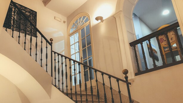 A staircase with ornate balusters and a handrail, leading to a landing with a large arched window and a wall-mounted light fixture.