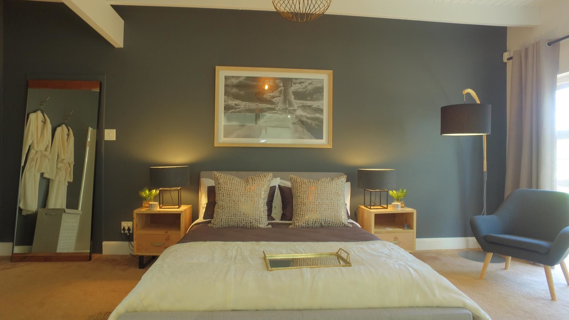 Ein modernes Schlafzimmer mit einem großen Bett, zwei Nachttischen, einem Sessel, einem Spiegel und einem gerahmten Kunstwerk an der Wand.