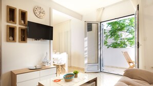 A modern living room with a flat-screen TV, wooden cabinets, a coffee table, and a sliding glass door leading to a balcony with a view of trees.