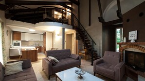 A spacious living area with a sofa, armchair, and a fireplace. A kitchen is visible in the background, and a staircase leads to an upper level.