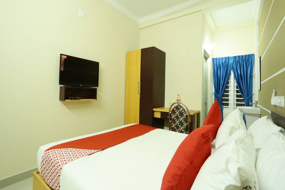 A hotel room with a bed, a TV mounted on the wall, a wardrobe, a desk with a chair, and a bathroom with a mirror and curtains.