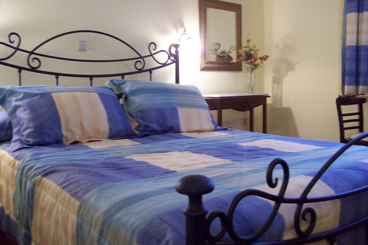 Un lit recouvert d’une couvre-lit à rayures bleues et blanches, une table de chevet en bois et un miroir accroché au mur.