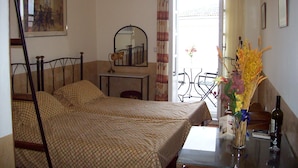 A bedroom with a bed, a bedside table, a mirror, a window with curtains, a small balcony with a table and chairs, and a vase with flowers.