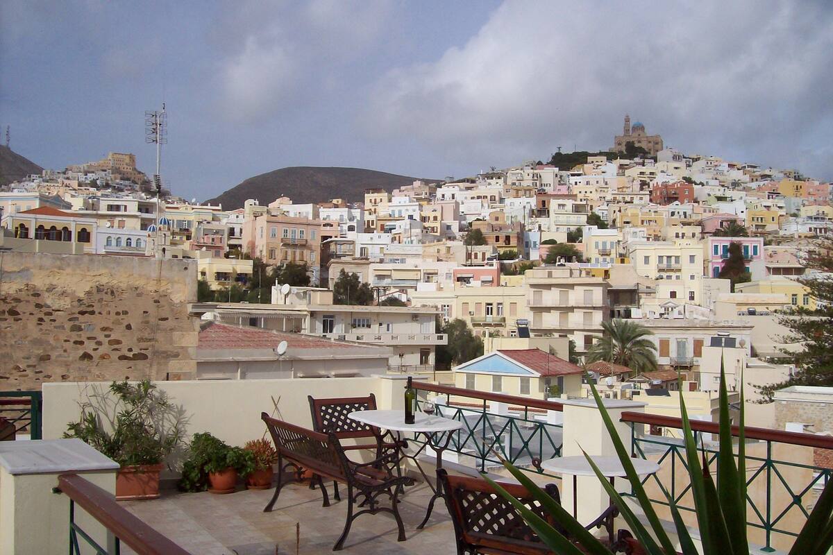 Une terrasse avec mobilier d’extérieur, une vue sur une ville pittoresque nichée sur une colline, et, au loin, une autre colline surmontée d’une structure ressemblant à un château.