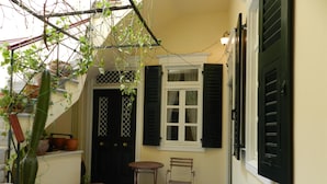 A small courtyard with a black door, a white window with shutters, a wooden chair, and potted plants.