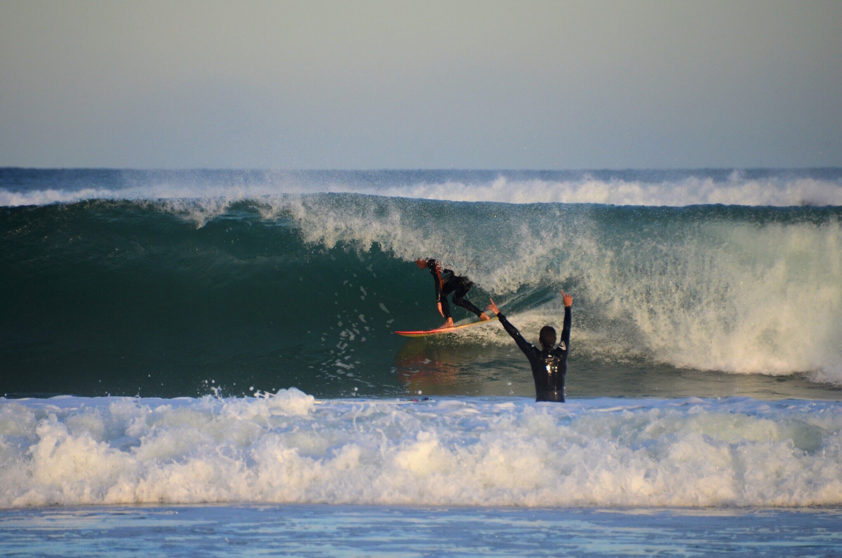 A surfer riding a large wave with another person watching from the shore.
