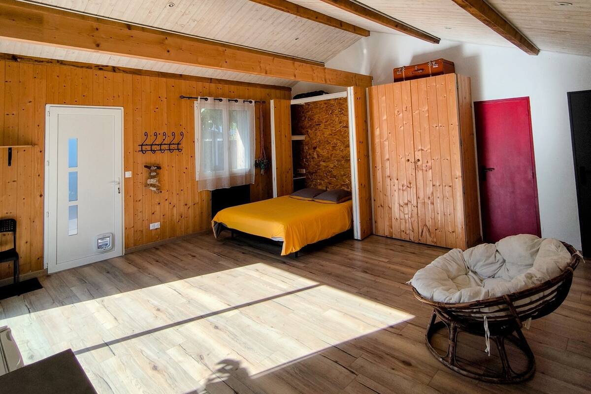 Una habitación con paredes y techo de madera, una cama con colcha amarilla, una silla de mimbre y una puerta roja.