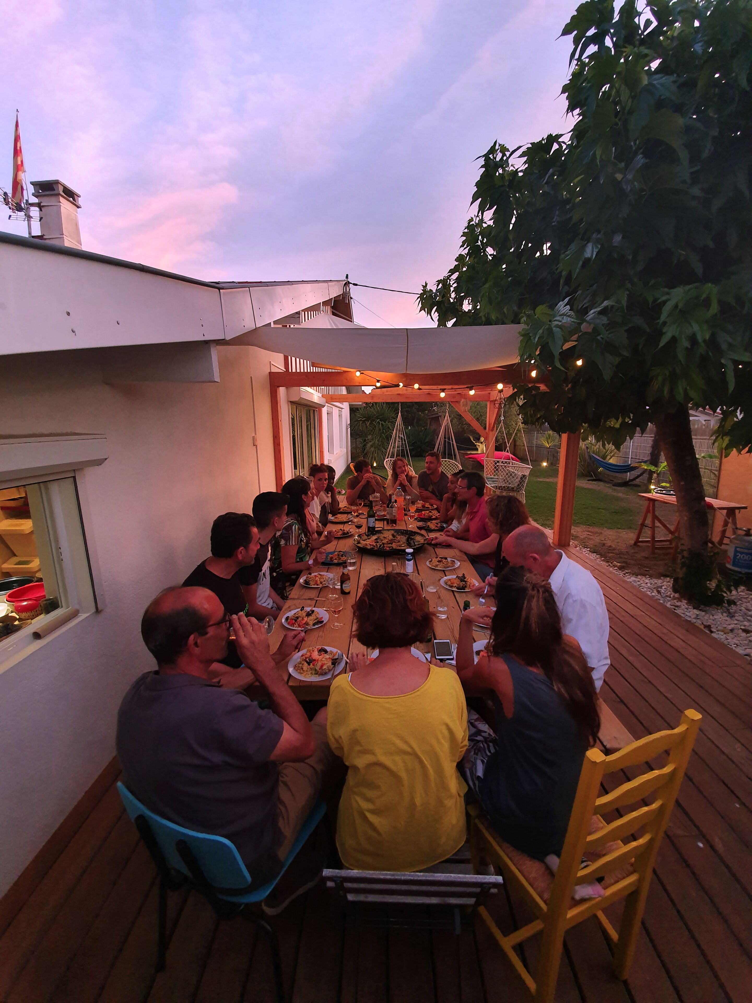 Un grupo de personas sentadas alrededor de una mesa de madera en un patio al aire libre, con una pérgola encima y luces colgantes.