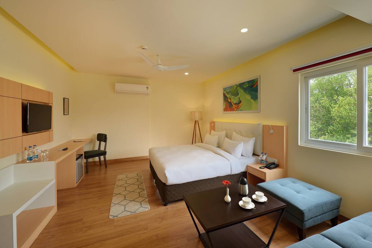 Une chambre d’hôtel comprenant un lit, une table de chevet, une chaise, un bureau, une télévision et une fenêtre donnant sur des arbres.
