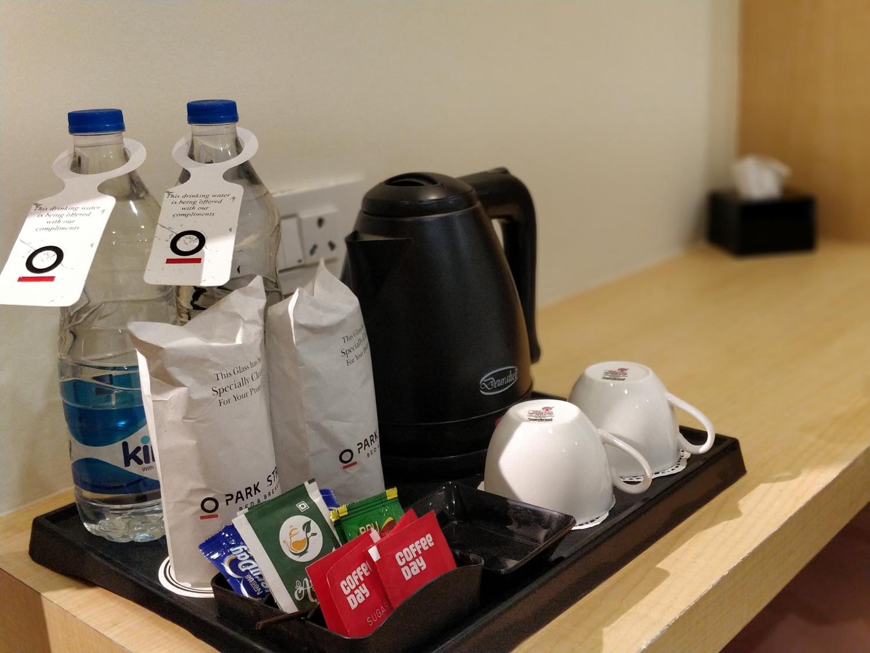 Hotellihuoneen aamiaistarjotin, jossa on kahvinkeitin, kaksi pulloa vettä, lasi, sokeripusseja ja kahvipussi.