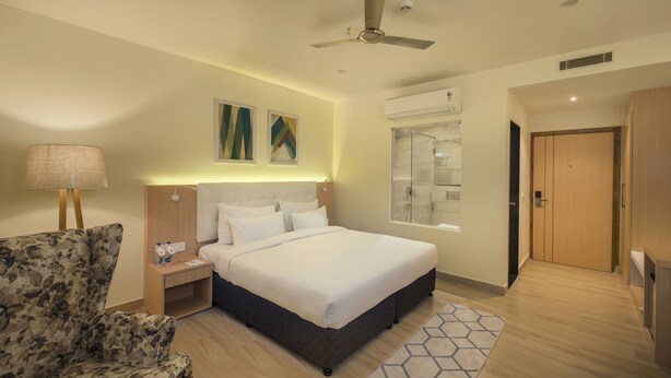 Une chambre d’hôtel avec un lit, une chaise, une table de chevet, une lampe, un ventilateur de plafond et une fenêtre.