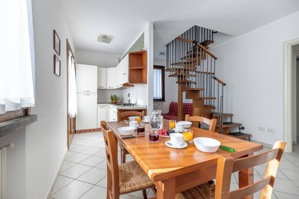A modern kitchen with white cabinets, a wooden dining table set with breakfast items, and a staircase with a wooden railing.