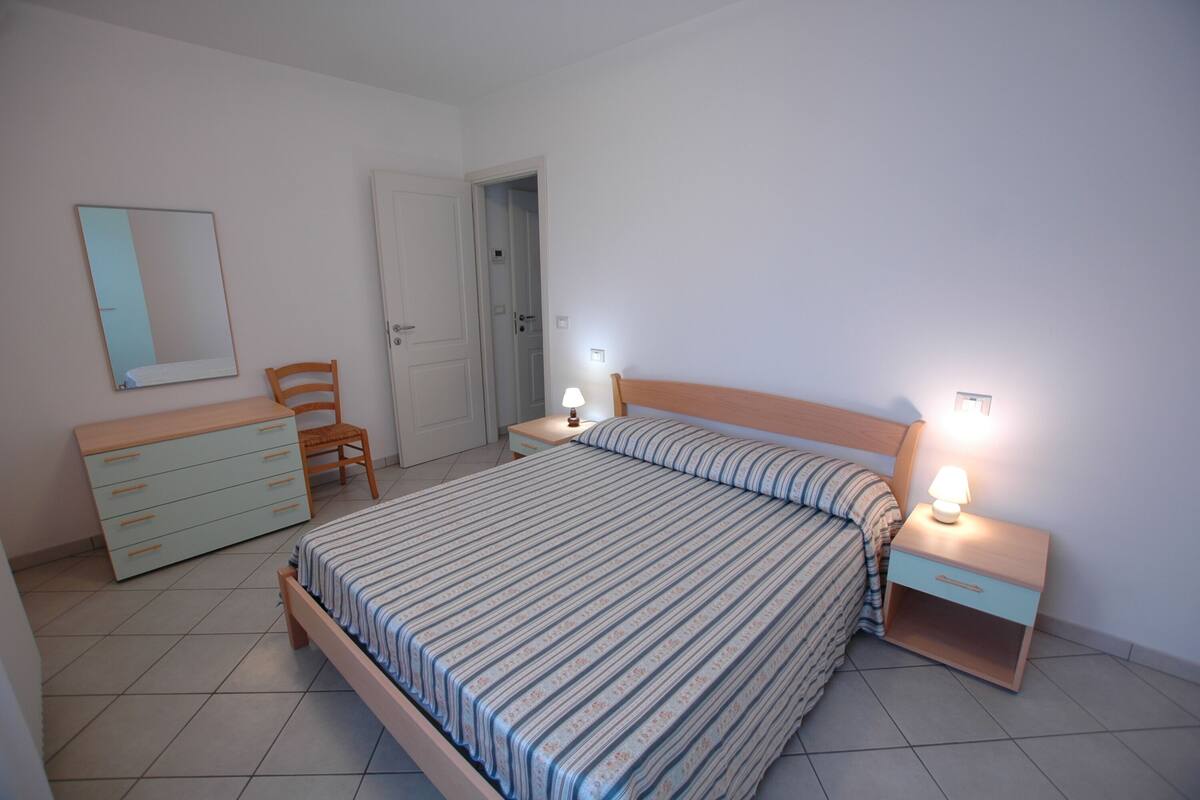 A bedroom with a bed, a nightstand with a lamp, a dresser, a chair, and a door leading to another room.
