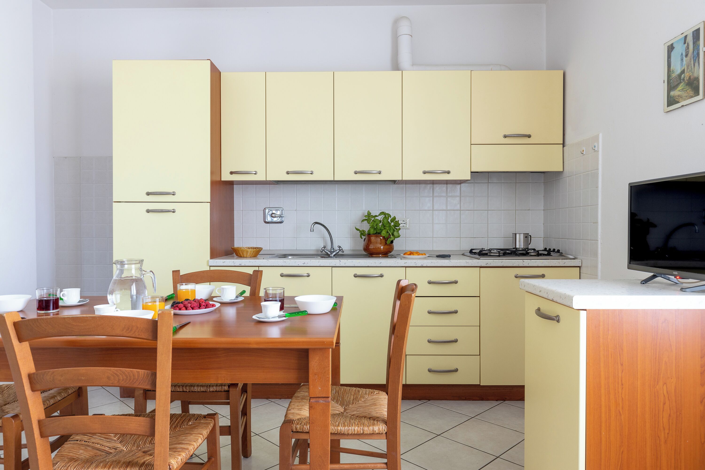 Una cucina con tavolo, sedie, pensili, lavello e televisore.