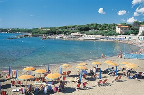 A beach with rows of sun loungers and umbrellas, a clear blue sea, and buildings in the background.