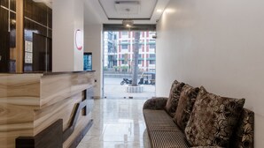 A modern reception area with a wooden reception desk, a sofa with patterned cushions, and a view of the street outside. - OYO 3551 Hotel Elite Inn (Navi Mumbai)