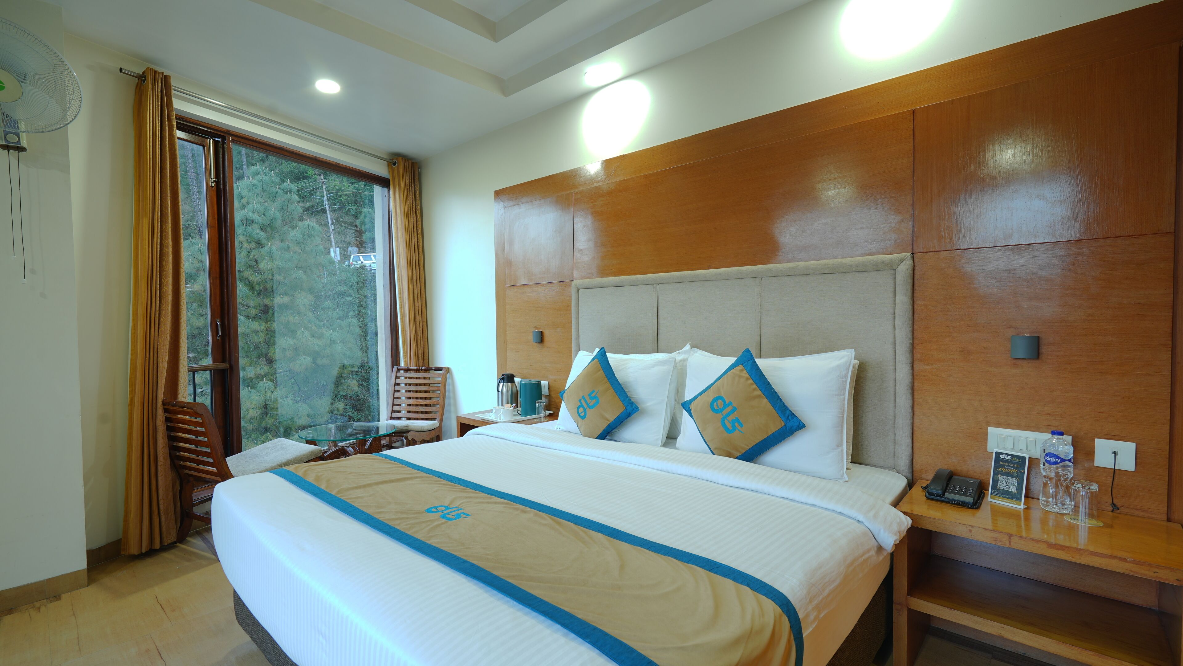 Quarto de hotel com uma cama grande, cabeceira de madeira, mesa de vidro, uma cadeira, mesa de cabeceira com um telefone e uma garrafa de água, e vista para árvores pela janela.