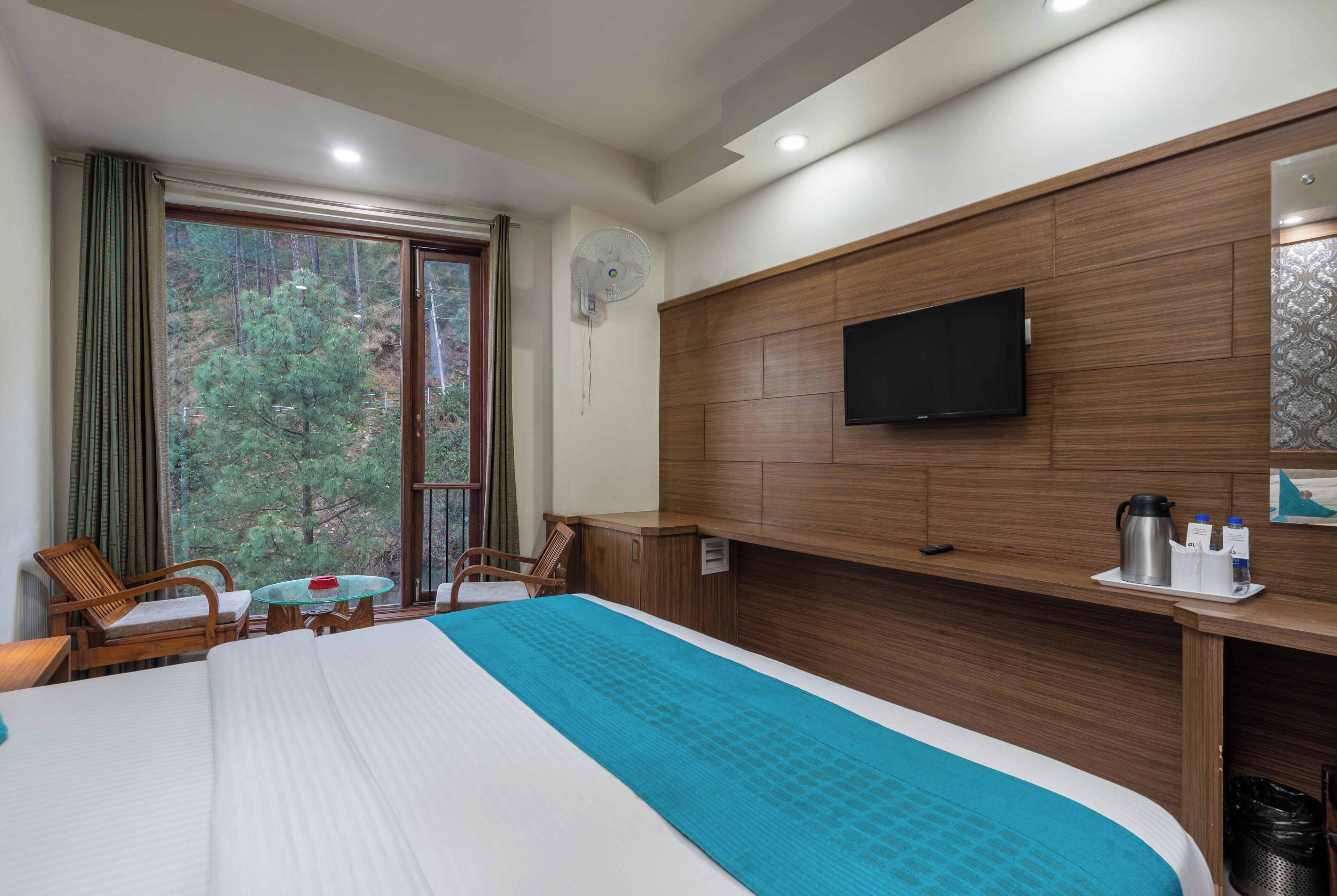 A hotel room with a large bed, a TV mounted on the wall, a desk with a coffee maker, and a window with a view of trees.
