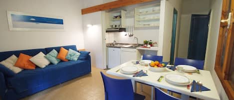A compact living space with a kitchenette, a dining table set for two, and a blue sofa with colorful cushions.
