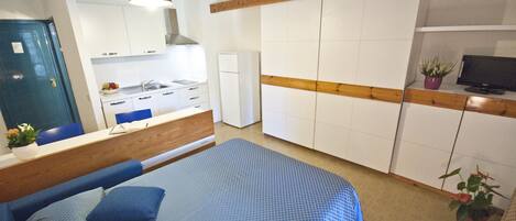 A bedroom with a bed, a wooden headboard, a bedside table, a nightstand, a TV, a dresser, a wardrobe, and a small kitchen area with a sink and a refrigerator.