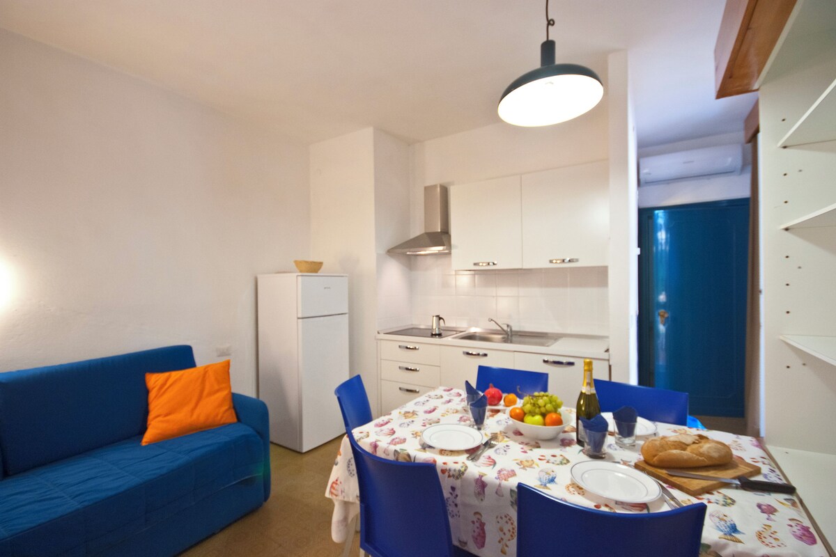 A compact kitchen with white cabinets, a blue sofa, and a dining table set with plates, a bottle, and a bowl of fruit.