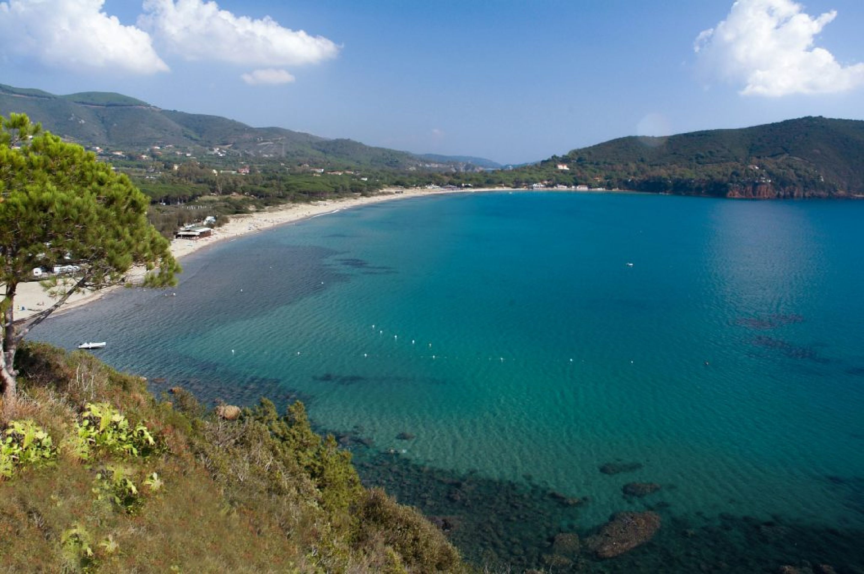 Un paisaje costero con mar azul transparente, playa de arena y una colina bordeada de árboles.