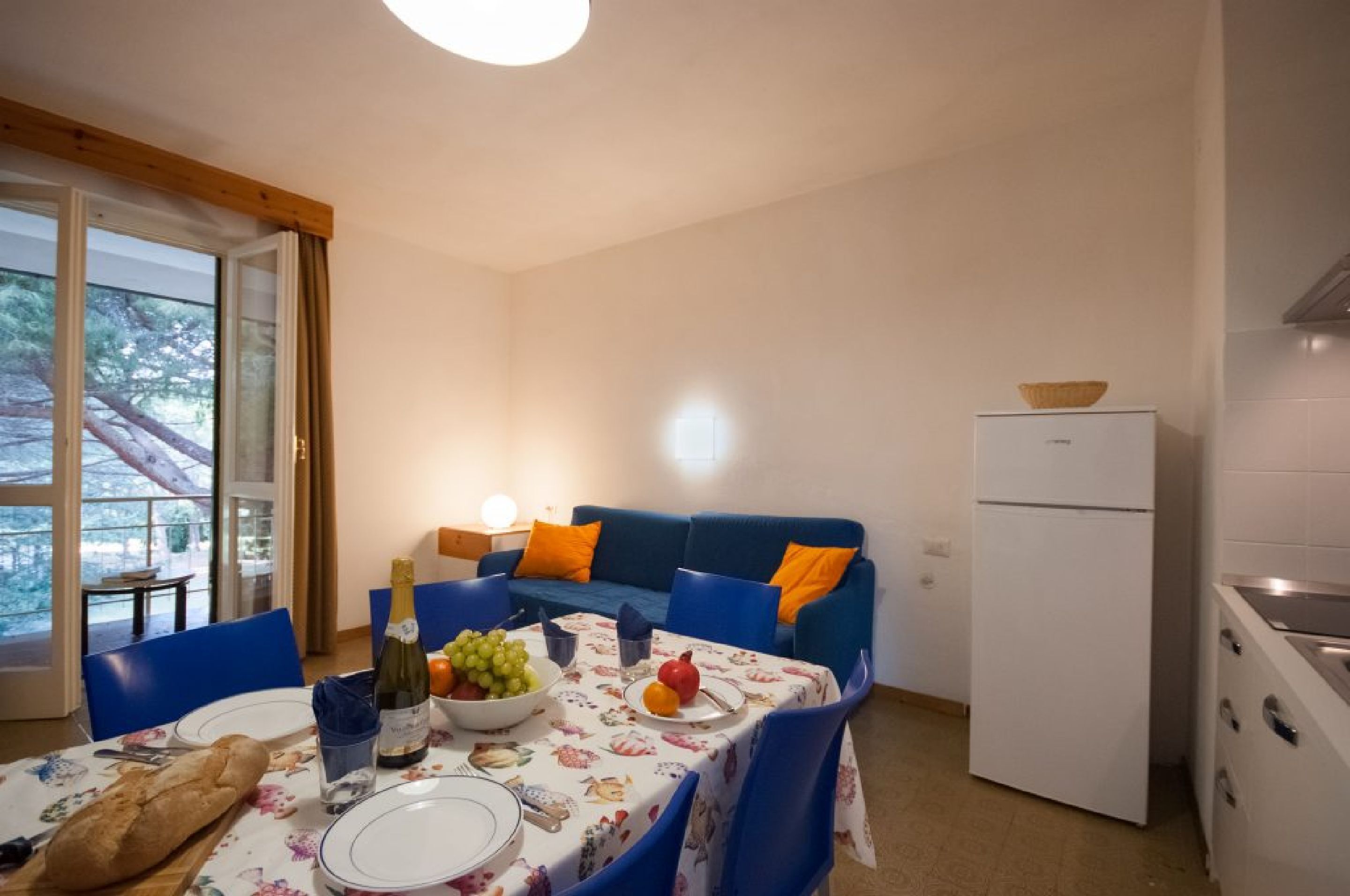 Un comedor con una mesa dispuesta para dos, un sofá azul con cojines naranjas, un refrigerador blanco y una ventana con vista a árboles.