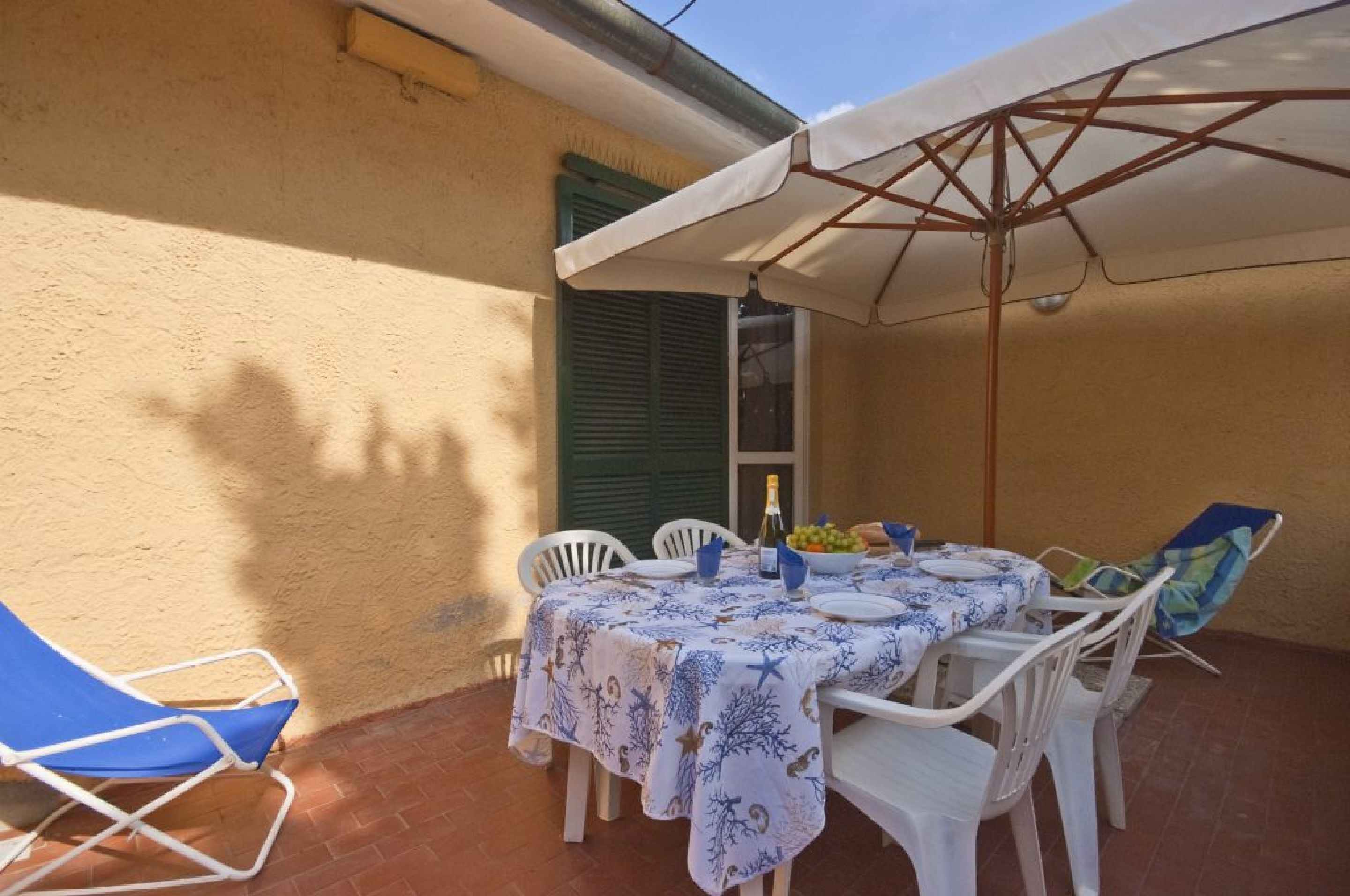 Un patio al aire libre con una mesa lista para comer, cubierta por un paraguas blanco y rodeada de sillas blancas.