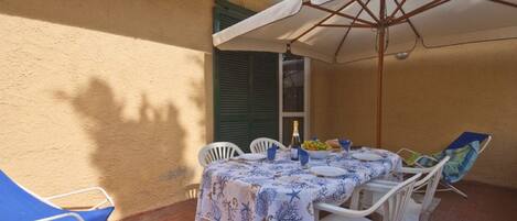Un patio al aire libre con una mesa lista para comer, cubierta por un paraguas blanco y rodeada de sillas blancas.