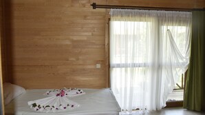 A wooden-paneled room with a bed, white curtains, and a view of greenery outside. - Serenity Bungalows (Kemer)