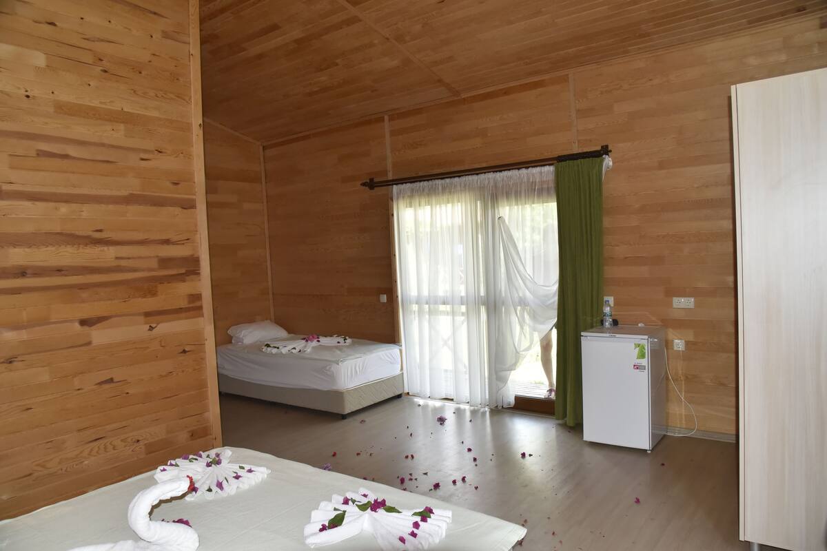 A wooden-paneled room with a bed, a small refrigerator, and a window with sheer curtains.