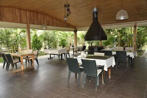 Un espacio para comer al aire libre con mesas y sillas de madera, rodeado de abundante vegetación.
