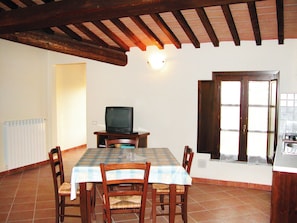 A dining area with a table set for four, a television on a stand, wooden chairs, and a kitchen area visible through a door. - Residence Giuly Rosselmini (Santa Luce)