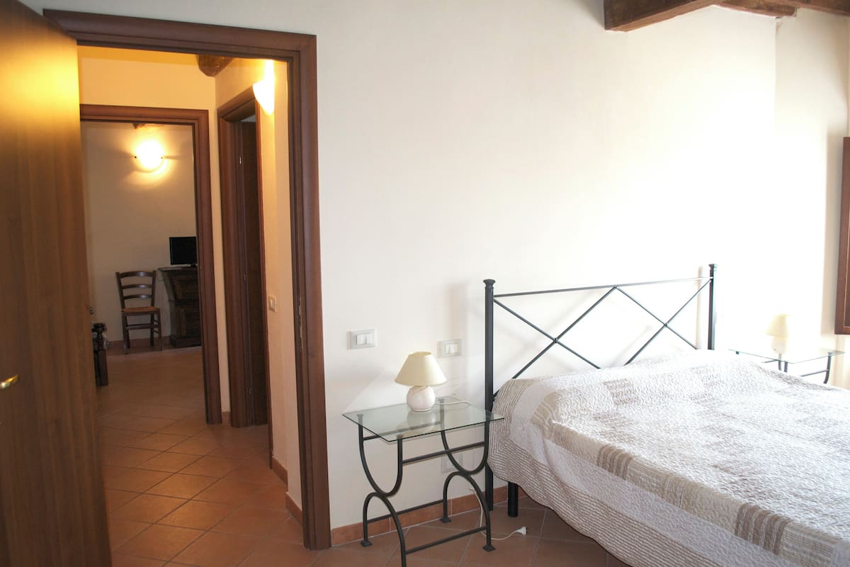 Un dormitorio con cama, una mesita de noche de cristal con lámpara, una silla al fondo y una puerta que da a otra habitación.