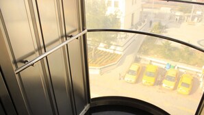 Une fenêtre incurvée, ornée d’une rambarde en métal, qui donne sur un parking où sont garées des voitures jaunes.