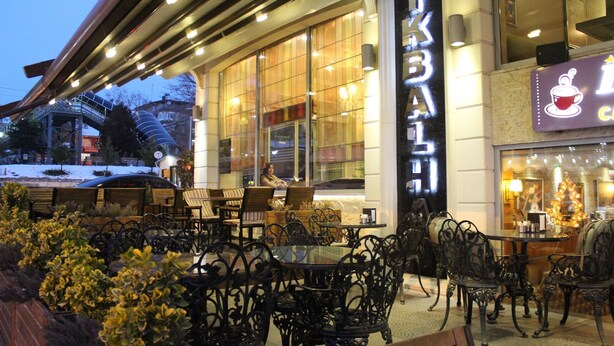 Un espace café en plein air avec des tables et des chaises, une grande fenêtre donnant sur la rue et une enseigne indiquant qu’il s’agit d’un café.