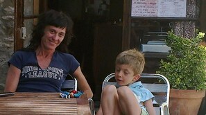 Een vrouw en een kind zitten aan een houten tafel buiten, met een potplant en een menubord op de achtergrond.