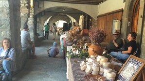 Un marché en plein air abrité, doté d’une arche en pierre, où une table expose différents types de fromages, et où l’on peut voir des gens assis et se promener.