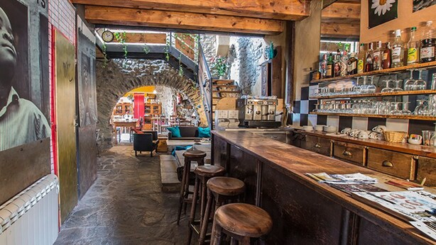 Un coin bar accueillant avec des tabourets de bar en bois, un comptoir où sont exposées diverses bouteilles et verres, et un mur en pierre rustique.