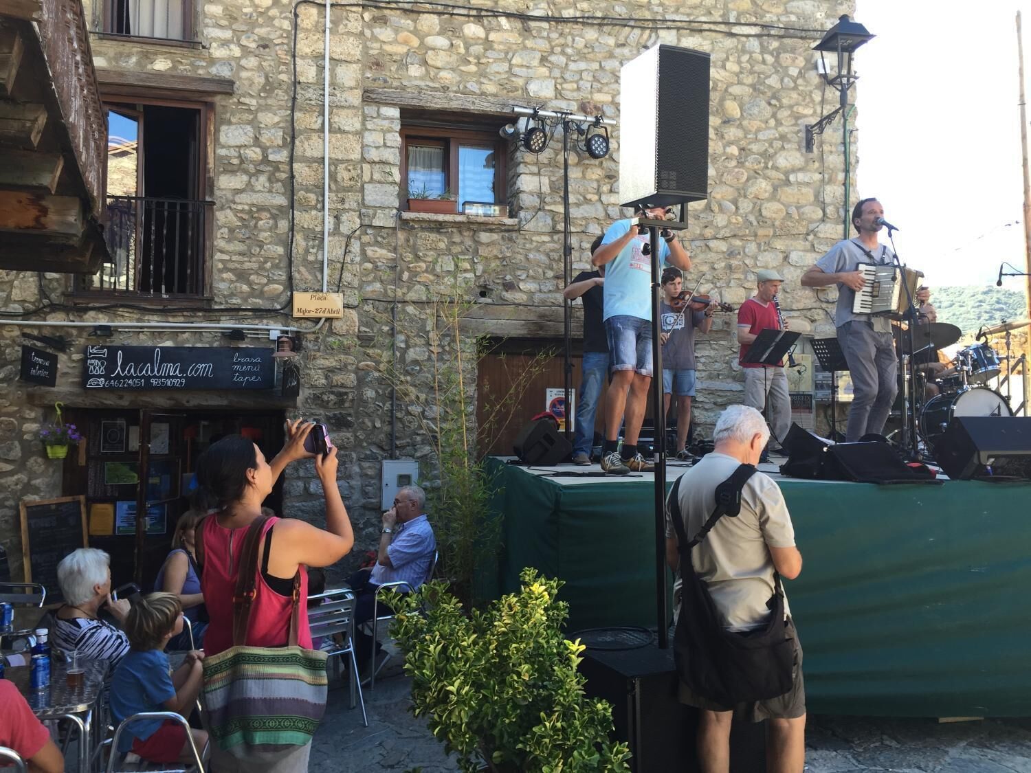 A street performance with musicians on a stage in front of a stone building with a cafe.