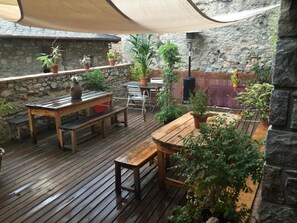 Una terraza de madera con una mesa y bancos, rodeada de plantas en macetas y un muro de piedra.